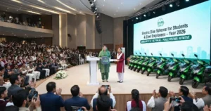An official ceremony in a large auditorium where a government representative hands a symbolic key to a recipient on stage as part of the Government of the Punjab Electric Bike Scheme 2026, with a row of green electric scooters parked nearby.